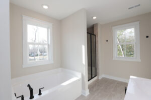 Modern bathroom in Poplar Village, Lot 1 with a white bathtub, glass-enclosed shower, and two windows letting in natural light.