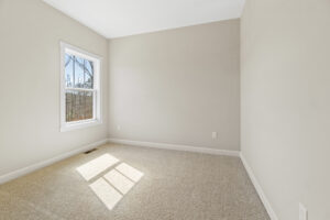 The empty room with beige walls in Poplar Village's Lot 1 offers a serene forest view through the window, where sunlight gracefully casts shadows on the carpeted floor.