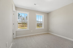 The empty room in Lot 1 features beige walls and two windows offering a serene view of trees, reminiscent of the tranquil landscapes in Poplar Village. The light-colored carpeted floor adds to the inviting ambiance, creating a peaceful retreat.