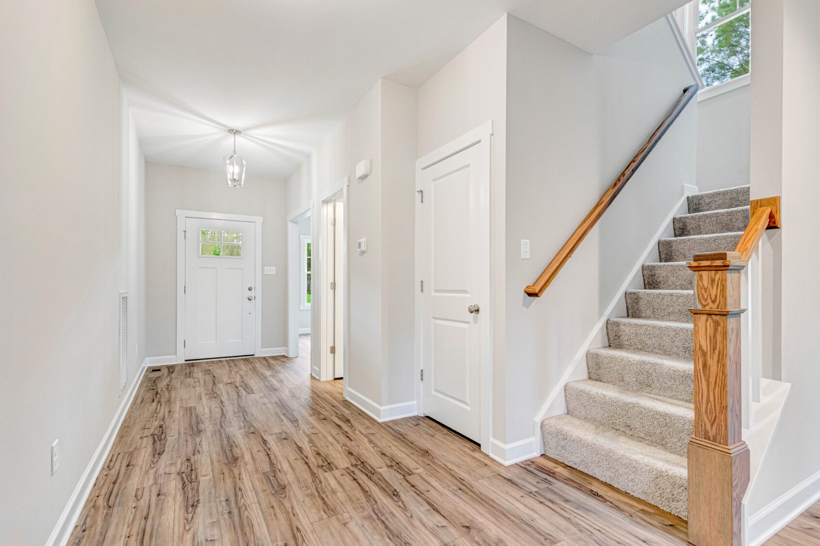 Bright hallway with wood floors, white walls, a staircase, and a front door with windows in the background.