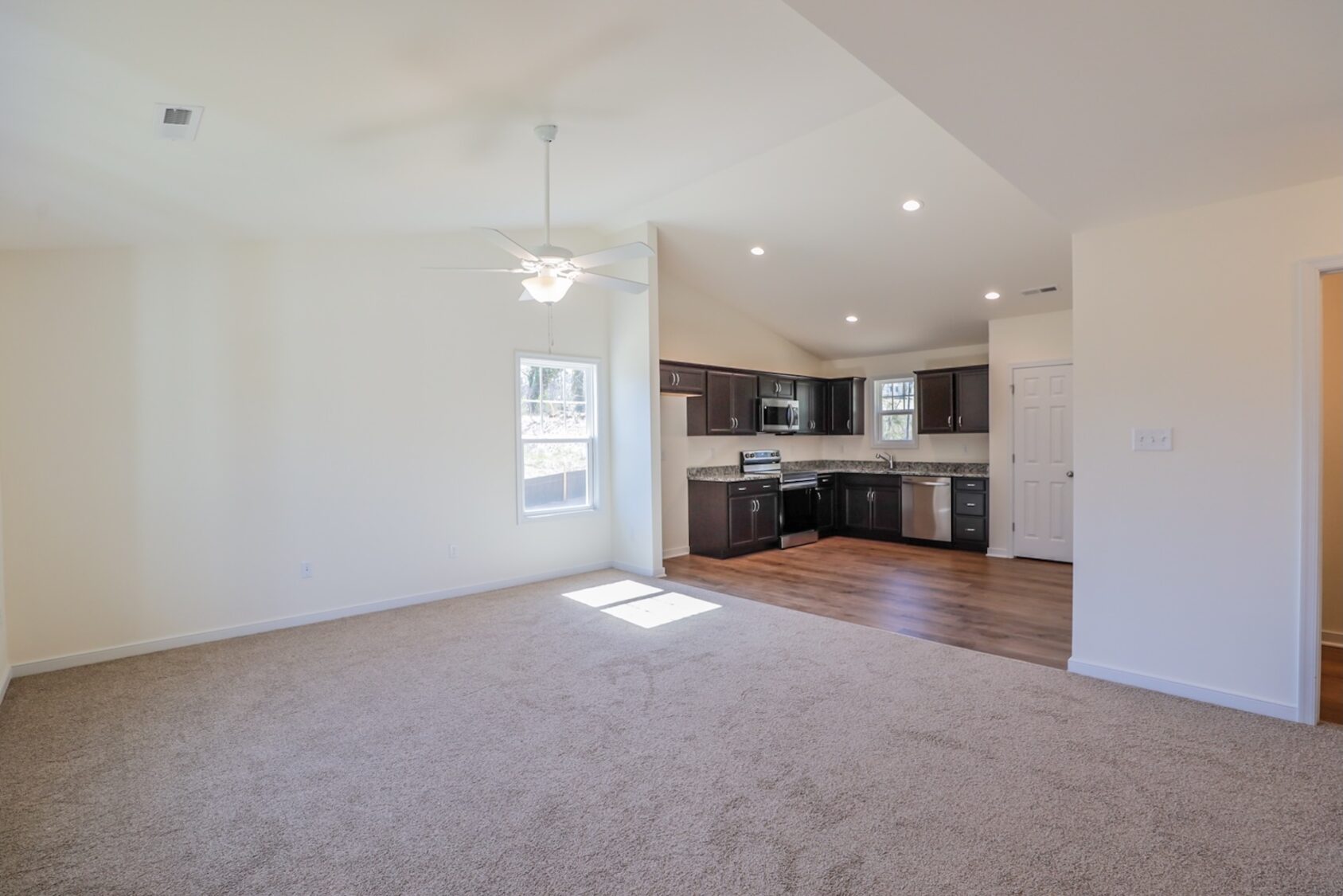 Open living room with beige carpet, ceiling fan, and view of modern kitchen with dark cabinets and wood flooring.