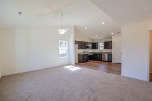 Open living room with beige carpet, ceiling fan, and view of modern kitchen with dark cabinets and wood flooring.