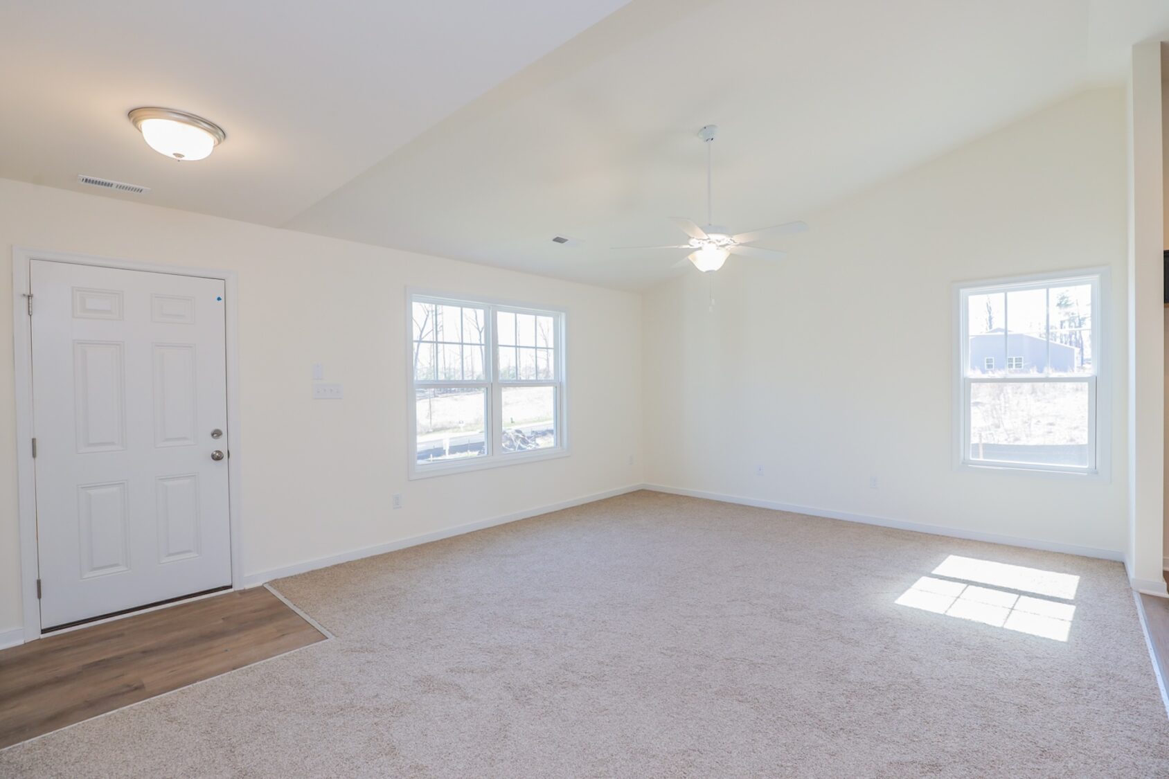 Bright, empty living room with beige carpet, white walls, ceiling fan, and large windows.