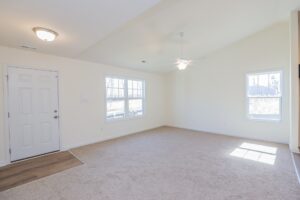 Bright, empty living room with beige carpet, white walls, ceiling fan, and large windows.