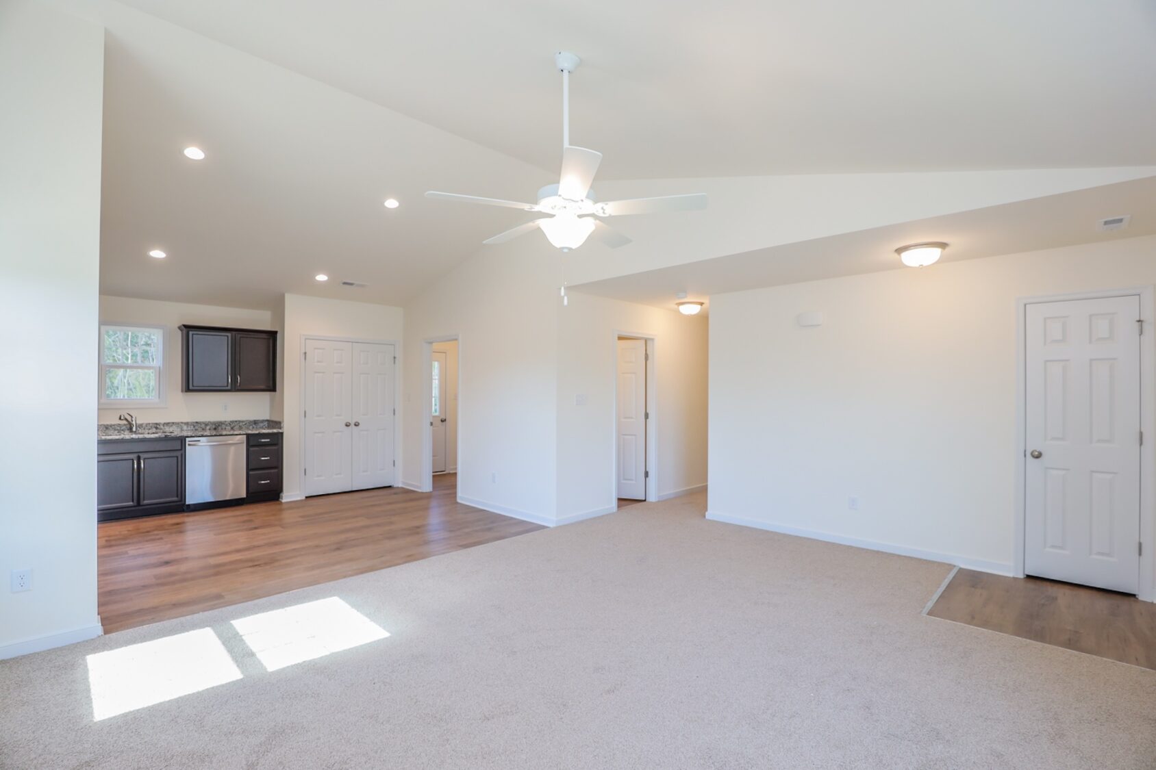 Bright open living area with carpet, ceiling fan, and view of kitchen with wood flooring and cabinets.