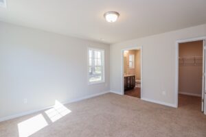 Bright empty bedroom with beige carpet, two windows, walk-in closet, and an open doorway to a bathroom.