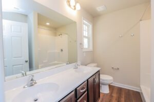 Modern bathroom with double sinks, large mirror, toilet, shower, beige walls, and wood-style flooring.