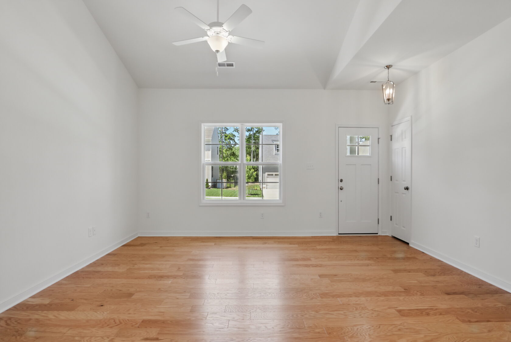 Bright empty room with light wood floors, white walls, ceiling fan, window, and front door with glass panes.