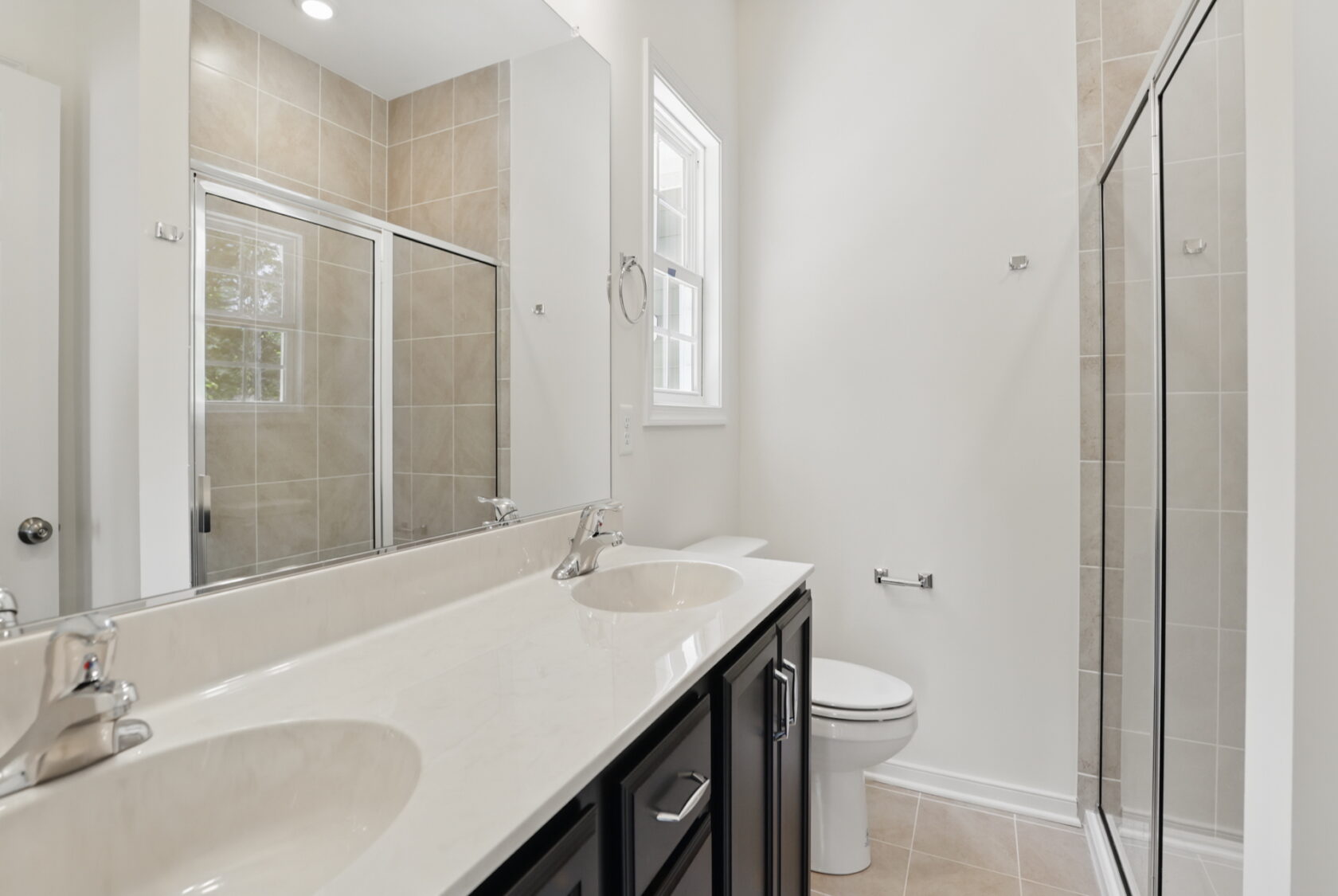 Modern bathroom with double sinks, a glass shower, toilet, large mirror, and beige tiled walls and floor.