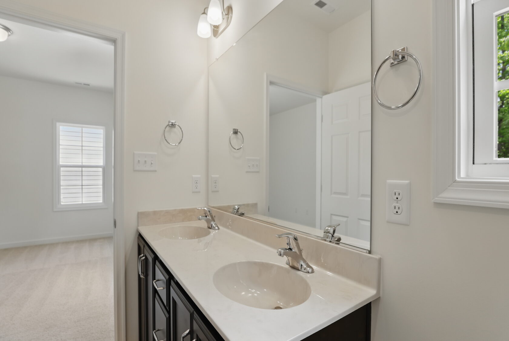 Bathroom with double sink vanity, large mirror, and view into an adjacent carpeted bedroom.