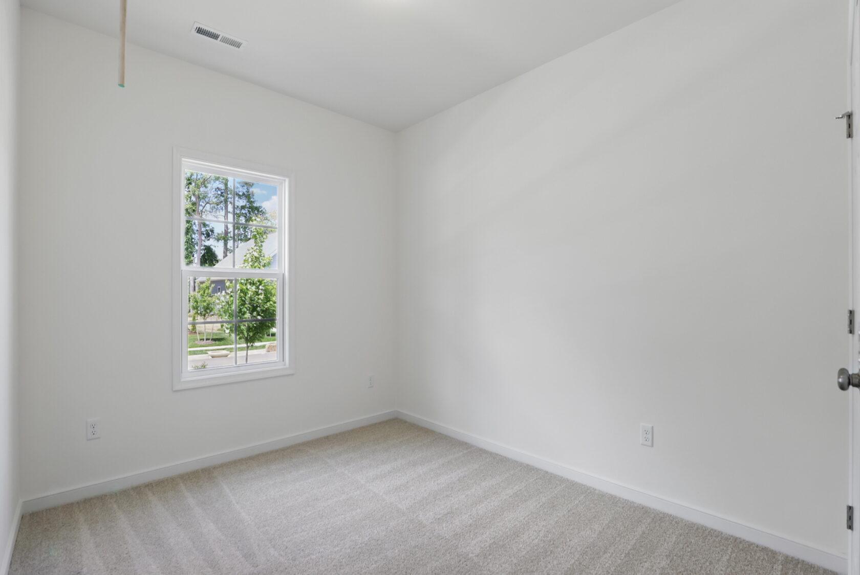 Empty room with white walls, beige carpet, and a window showing trees and blue sky outside.
