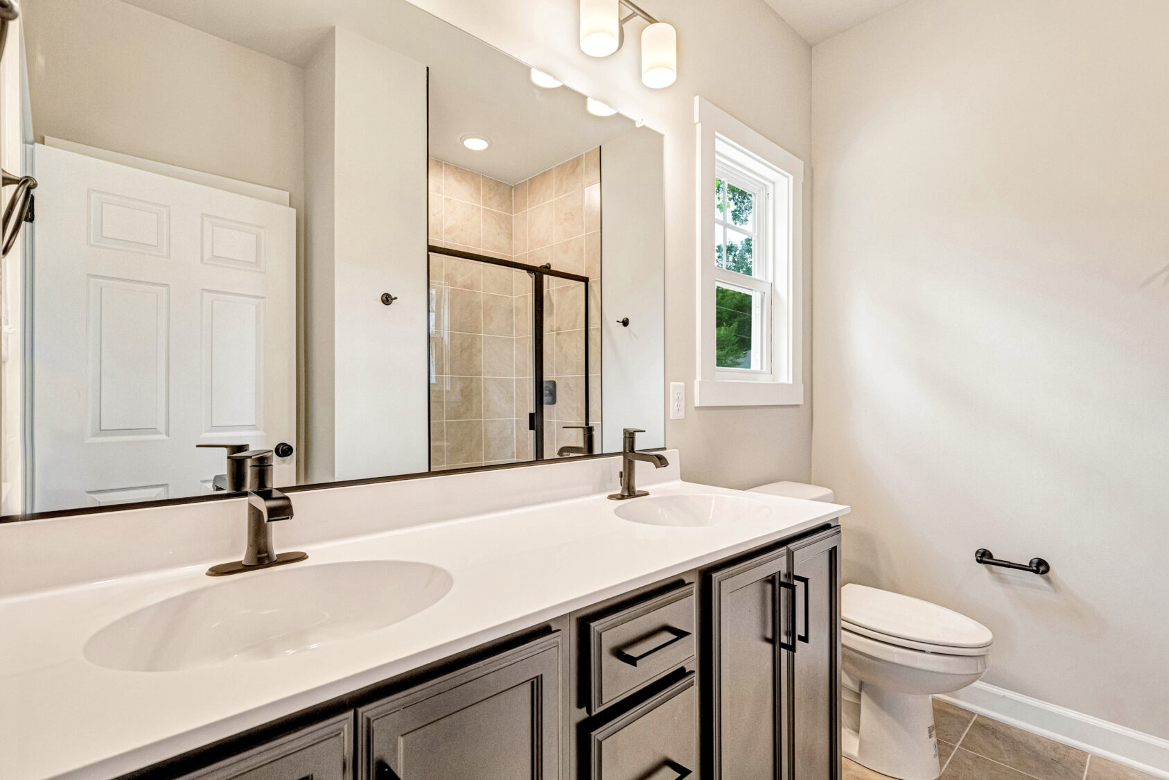 Modern bathroom with double sinks, black fixtures, a glass shower, white walls, and a window above the toilet.