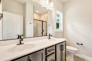Modern bathroom with double sinks, black fixtures, a glass shower, white walls, and a window above the toilet.