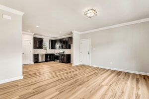 Bright, empty room with light wood floors, white walls, and a modern kitchen with dark cabinets in the background.