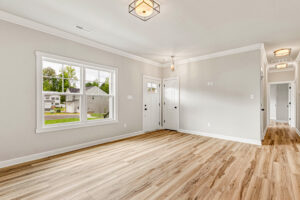 Bright, empty living room with wood floors, large window, and white walls, leading to a hallway and front door.