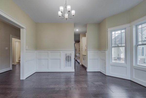Empty dining room with dark wood floors, beige walls, white wainscoting, and chandelier. Large windows on the right.