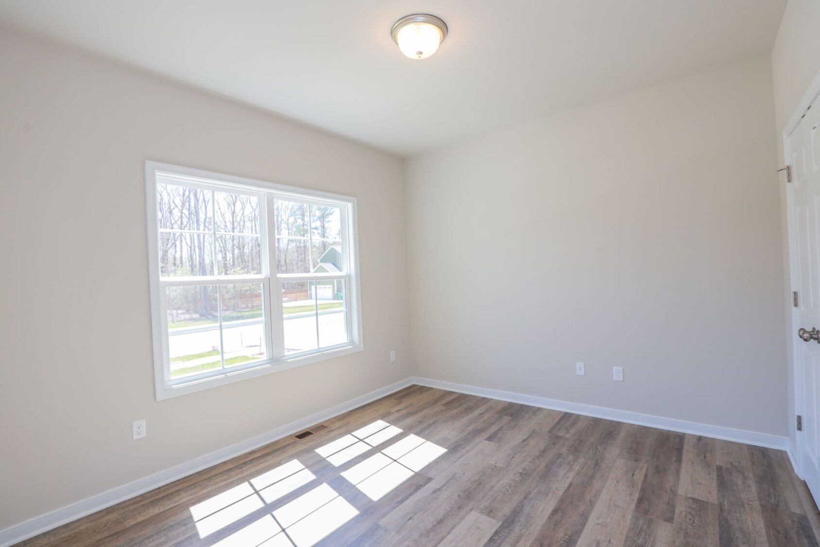 Empty room with light wooden floor, large window, white walls, and a ceiling light. Natural light fills the space.