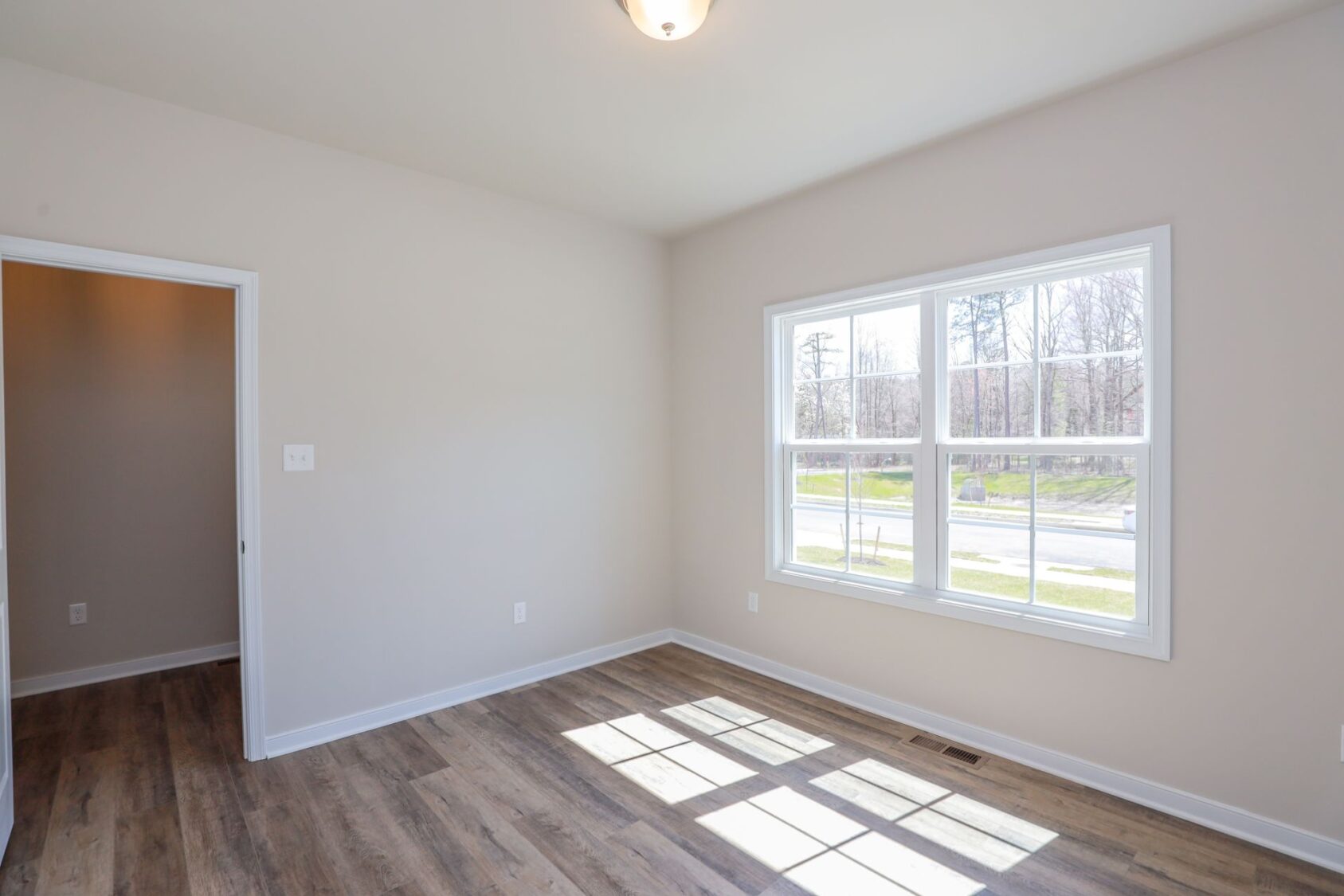 Empty room with a large window, wooden floor, and neutral walls, illuminated by natural light. Doorway visible on the left.