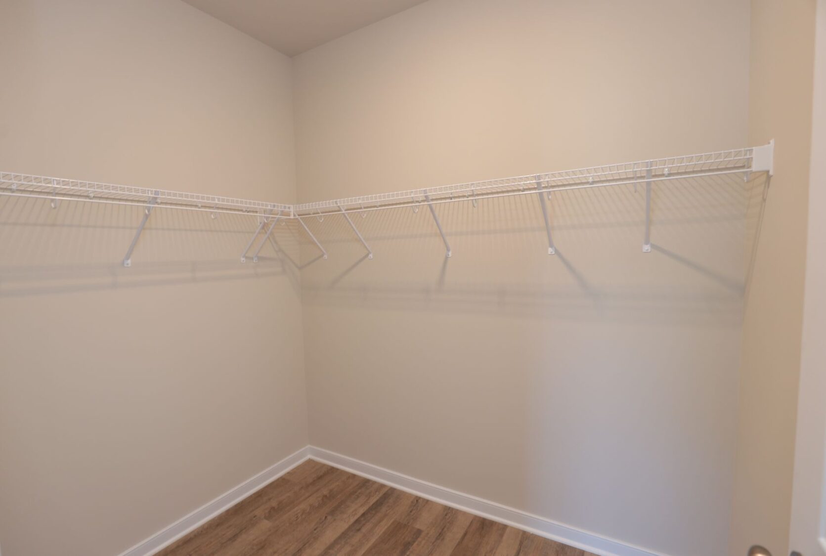 Empty walk-in closet with white wire shelving on beige walls and wooden floor.