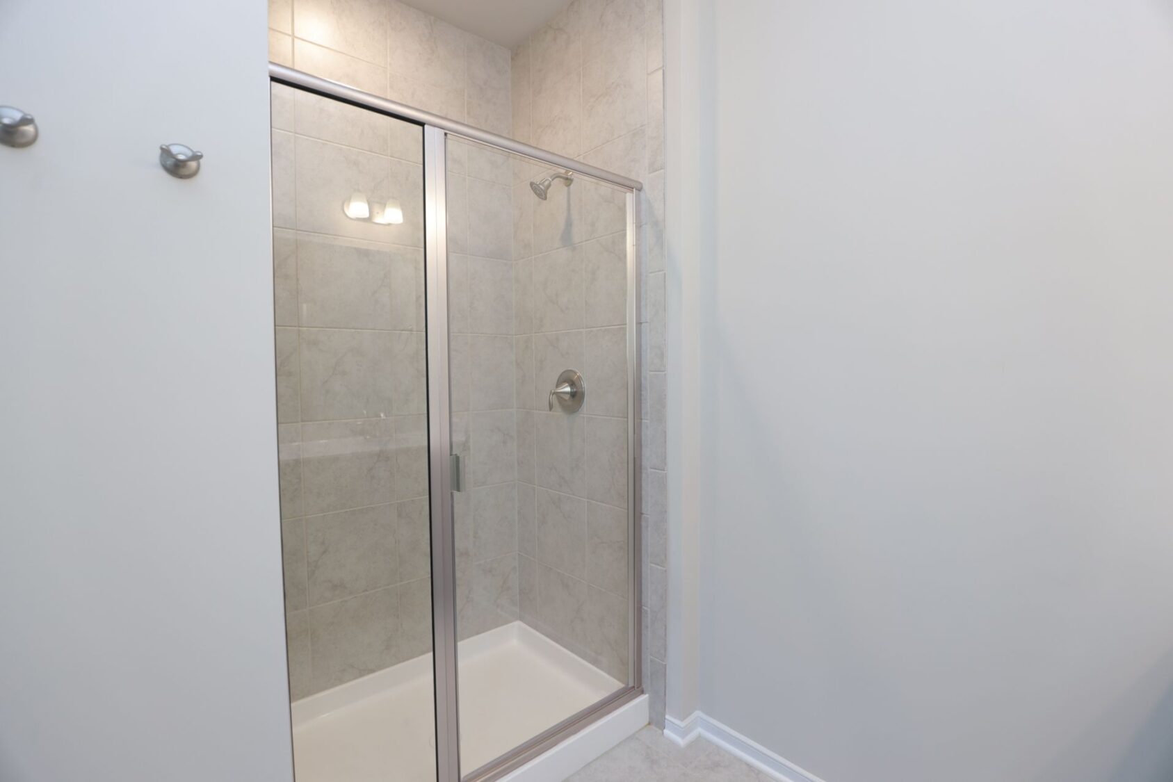 Glass shower stall with beige tiles, silver handle, and overhead light fixture in a bathroom.