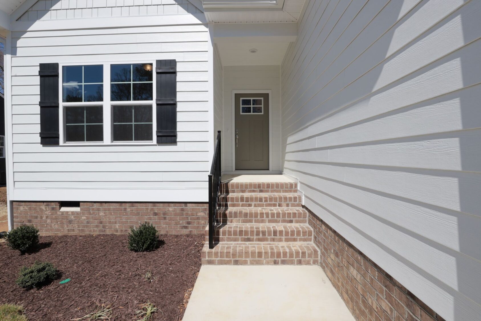 White house entrance with brick steps, black shutters, and a green door. Freshly landscaped with small bushes.