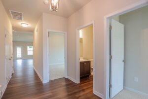 Hallway with wood floors, open doors to rooms and bathroom, and bright natural light from windows at the end.