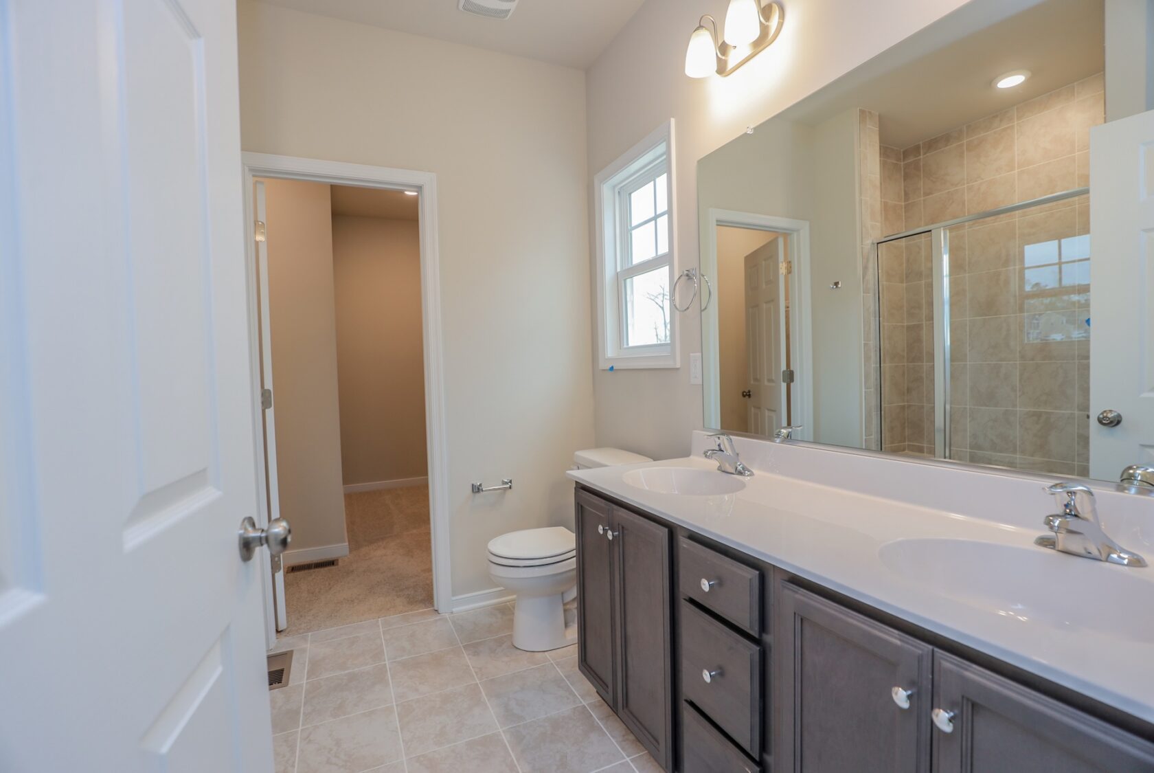 Modern bathroom with double sinks, a large mirror, toilet, window, and glass shower with beige tiles.