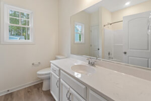 A bright bathroom in Lot 57 with a large mirror, white cabinets, sink, toilet, shower, window, and wood floor.