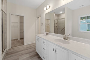 Modern bathroom in Lot 57 with dual sinks, large mirror, and walk-in closet. Neutral tones and wood flooring.