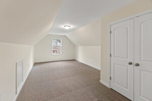 Empty attic room with beige walls, carpeted floor, white double doors, and a view of Patrick Henry Heights.