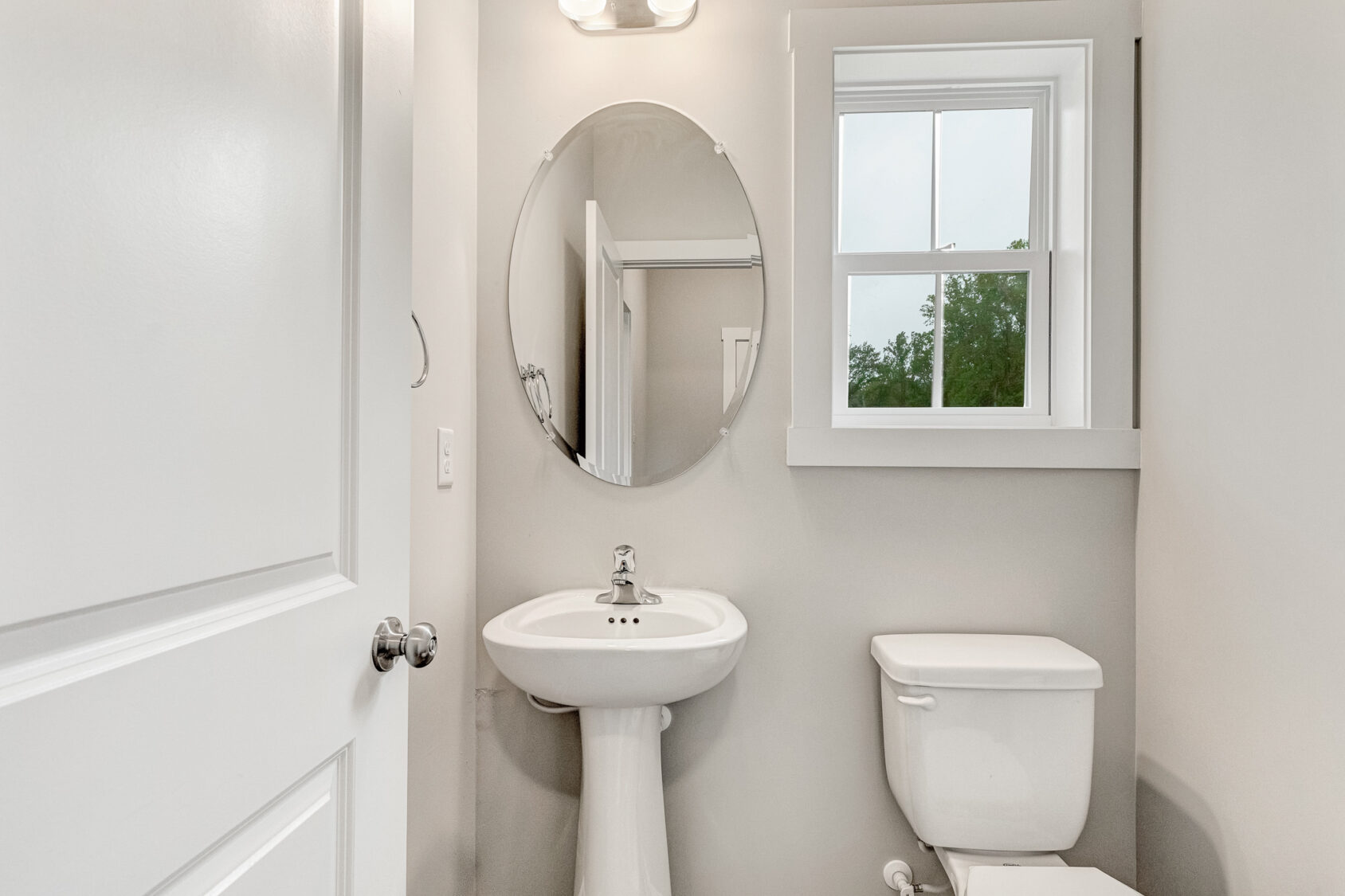Small bathroom in Lot 22, Poplar Village, with a pedestal sink, round mirror, toilet, and natural light from a window.