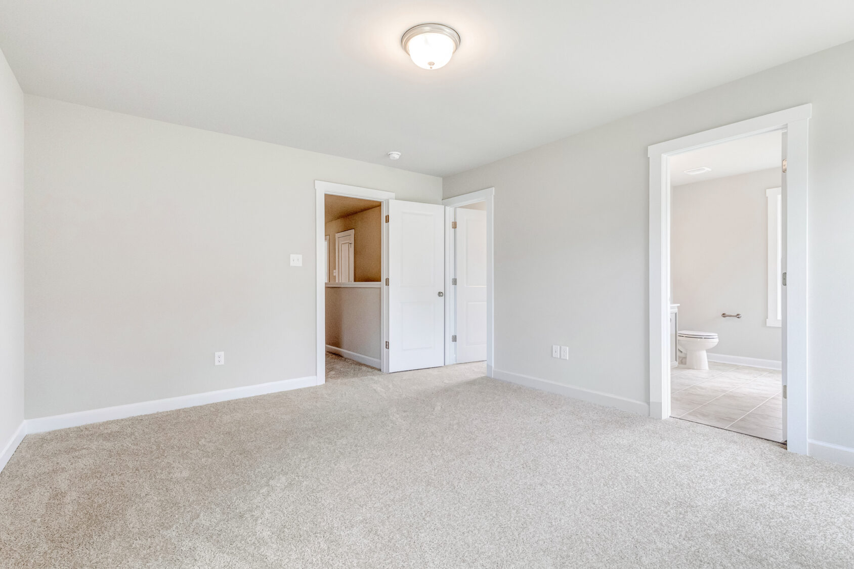 Bright, empty bedroom in Poplar Village with beige carpet, white walls, and a doorway leading to a bathroom.
