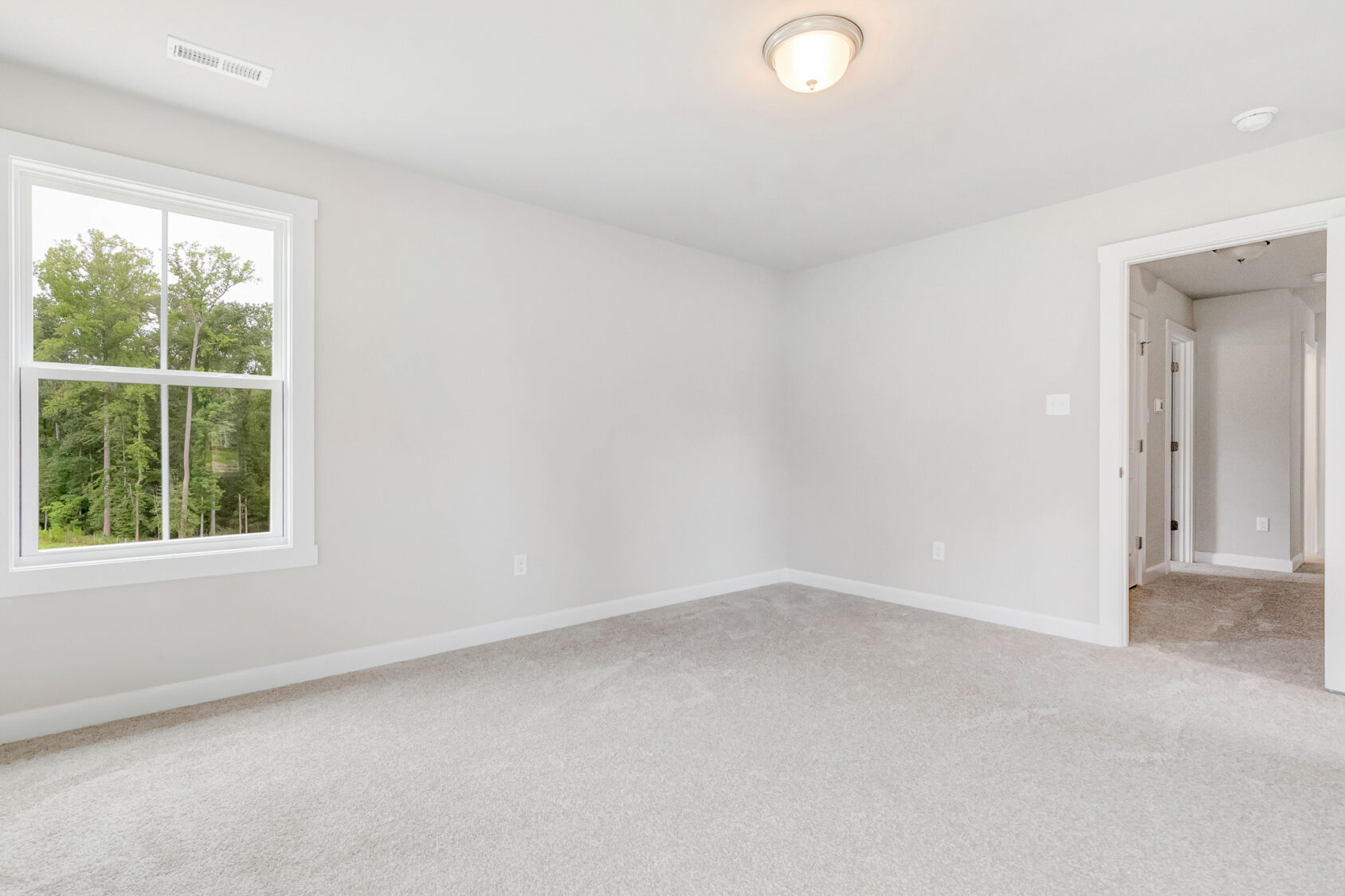 Empty, carpeted room in Poplar Village Lot 22 with white walls, a window showing greenery, and an open hallway doorway.