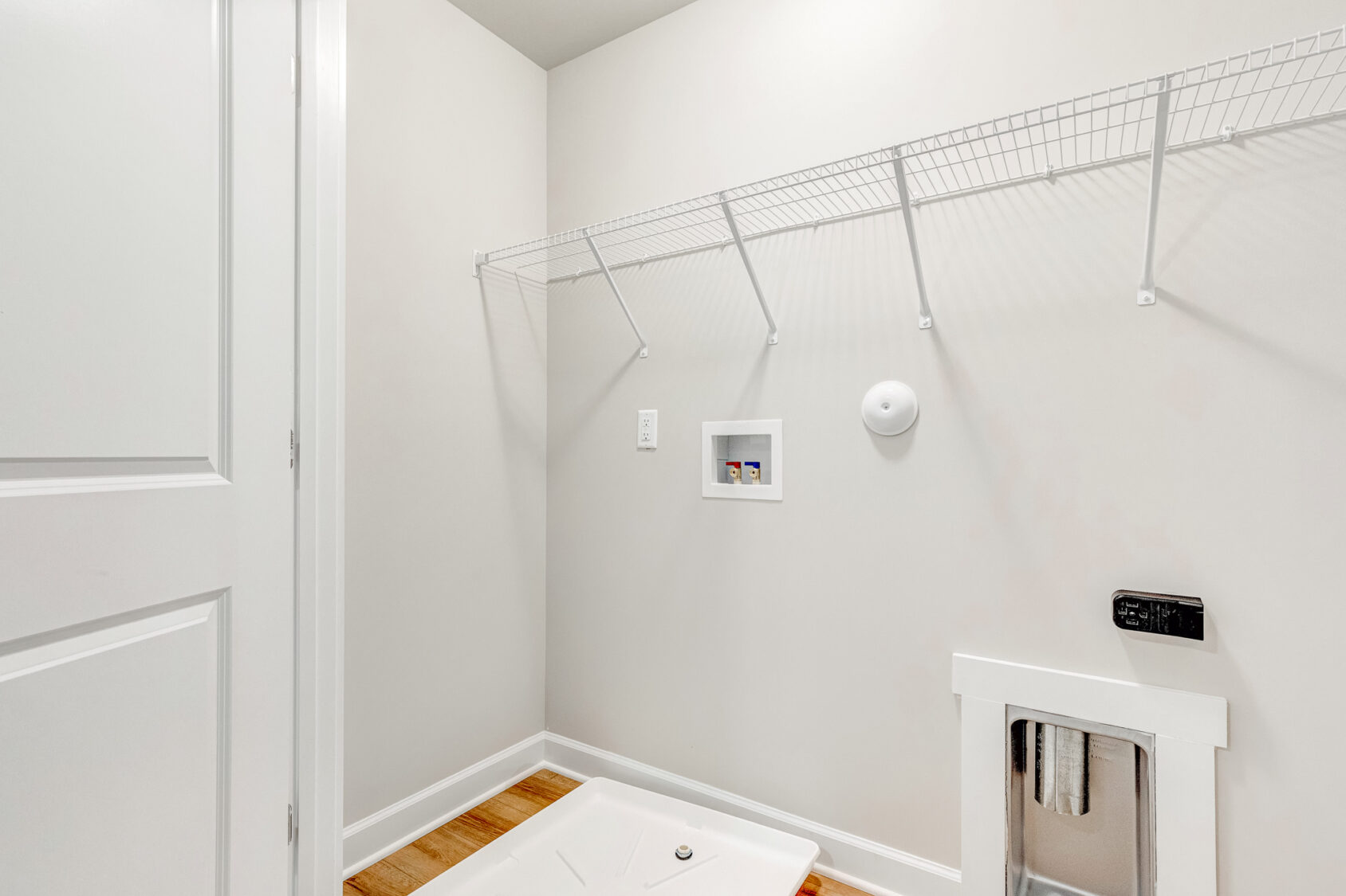 Empty laundry room in Poplar Villages Lot 22 with white walls, wire shelf, and hookups on a wooden floor.