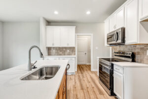 Modern kitchen at Poplar Village Lot 22 with white cabinets, stainless appliances, island sink, and light wood flooring.
