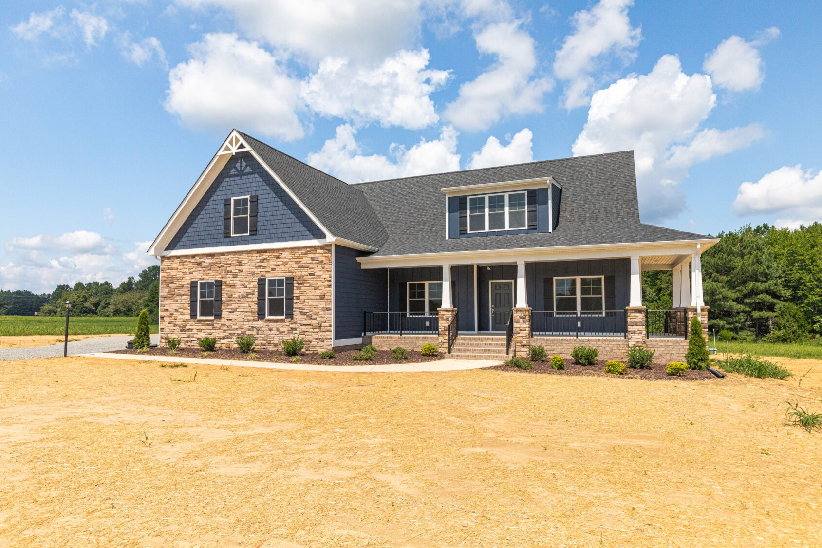 Modern two-story house with blue siding, stone accents, and a covered front porch, set on a sunny, open lot.