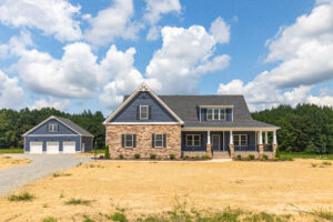 Modern house with stone and blue siding, three-car garage, and a large yard under a partly cloudy sky.
