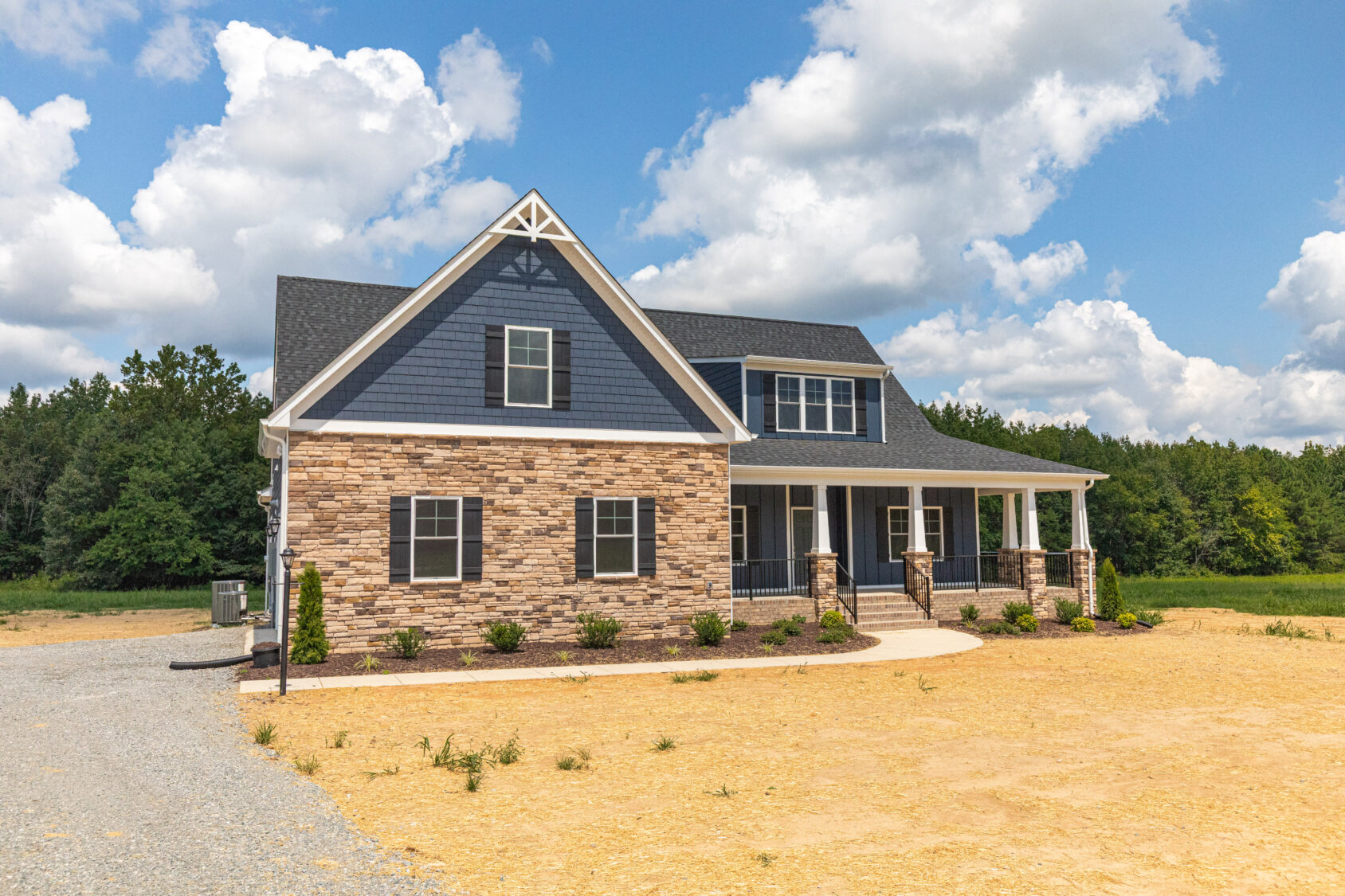 Two-story house with stone and blue siding, front porch, and gable roof on a large sandy lot.