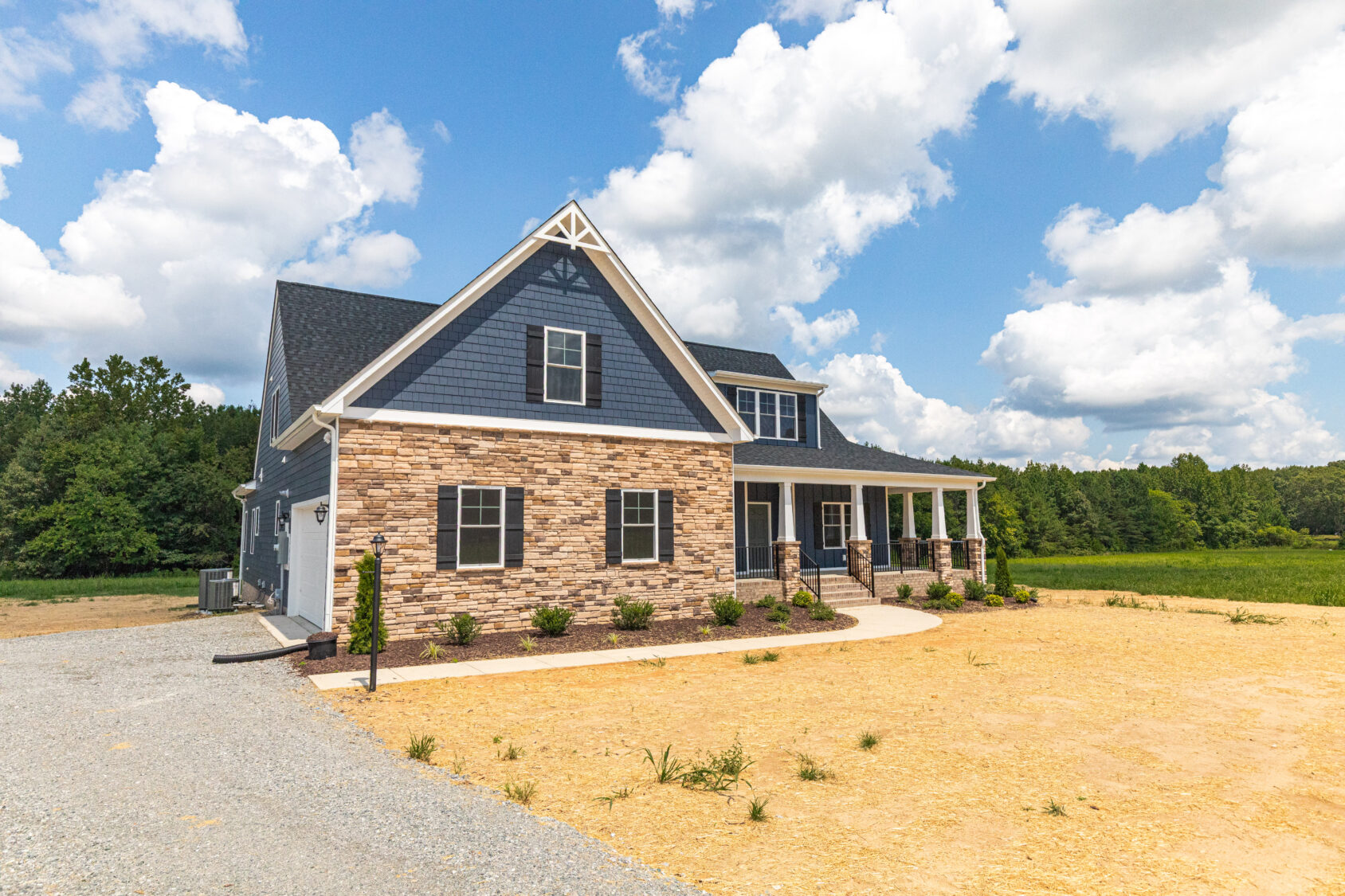 Two-story house with stone and blue siding, large porch, and gravel driveway under a partly cloudy sky.