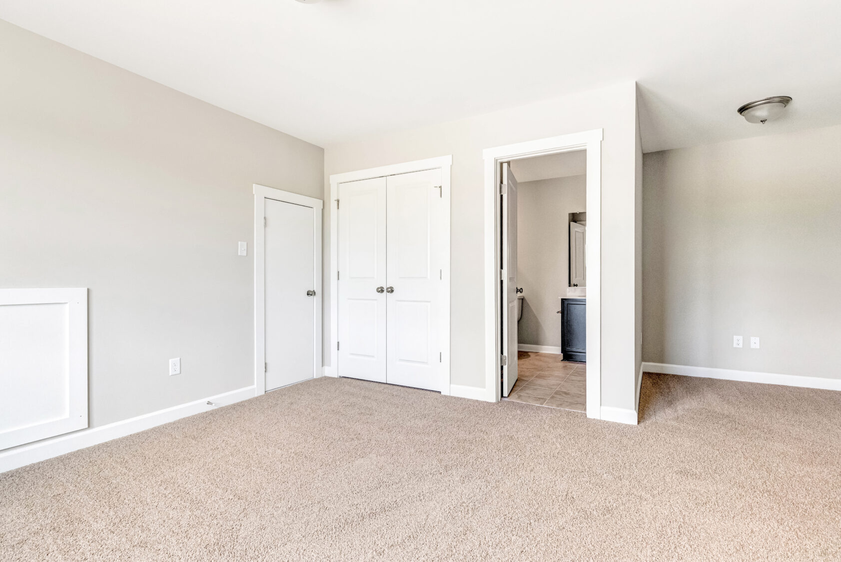 Bright, empty bedroom with beige carpet, white doors, and an open doorway leading to a bathroom.
