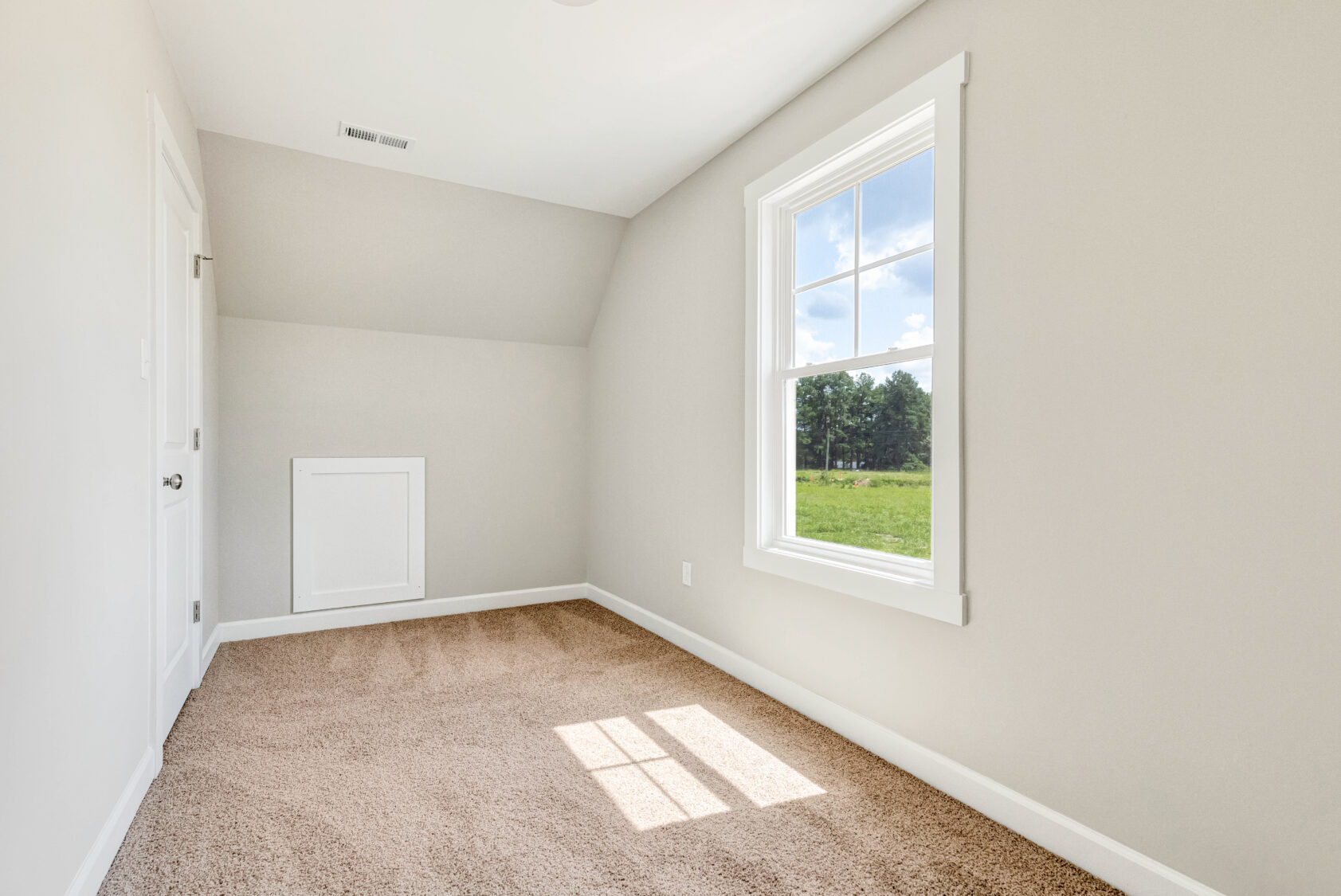 Small empty room with beige carpet, white walls, and a window showing a green field and blue sky outside.