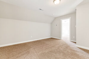 Empty carpeted room with beige walls and a ceiling light, open doorway leads to a similar adjacent space.