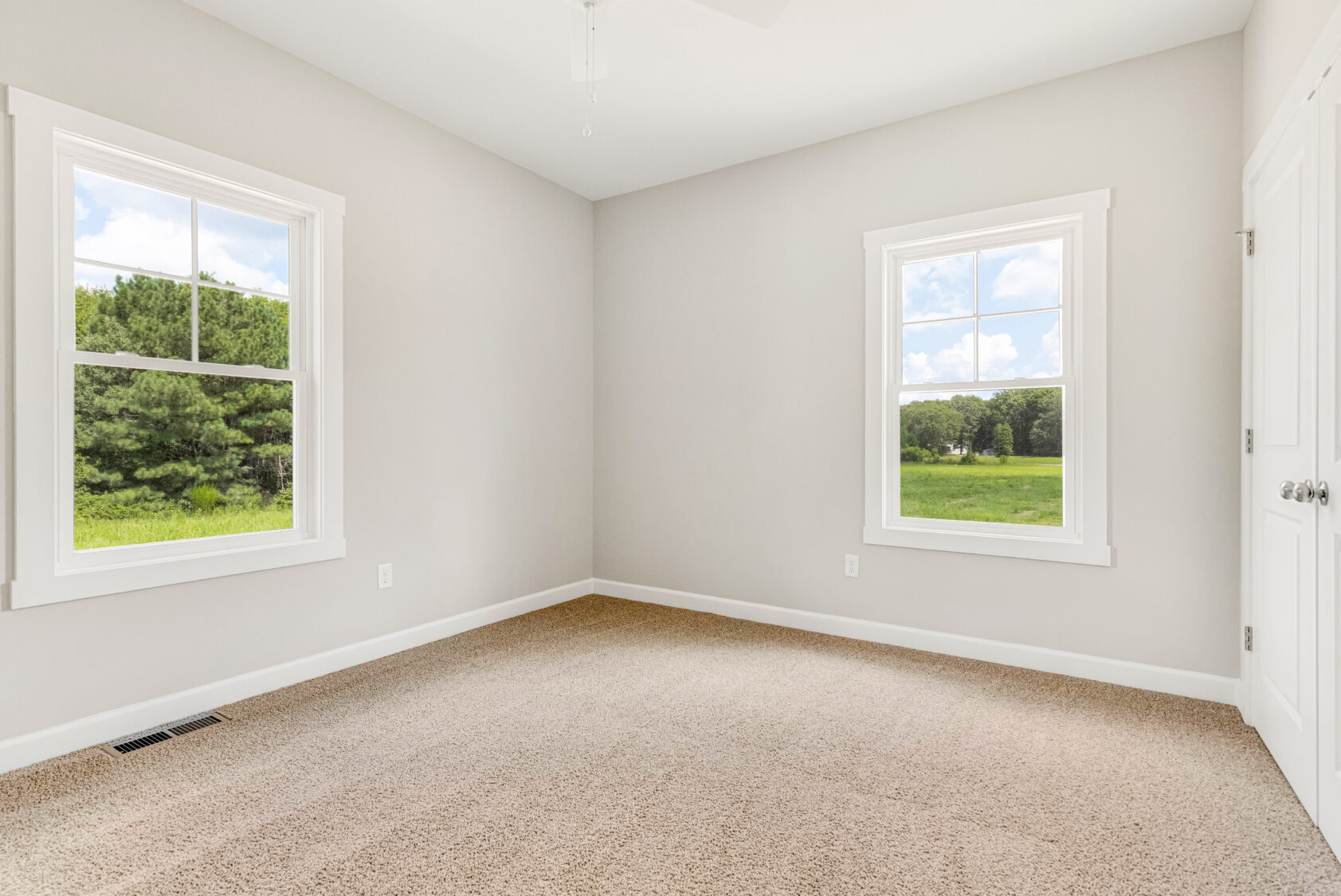 Empty room with beige carpet, two windows, white walls, and a view of greenery outside.