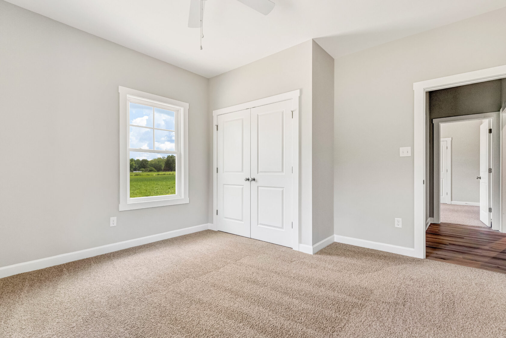 Bright, empty bedroom with beige carpet, white closet doors, and a window showing green fields outside.