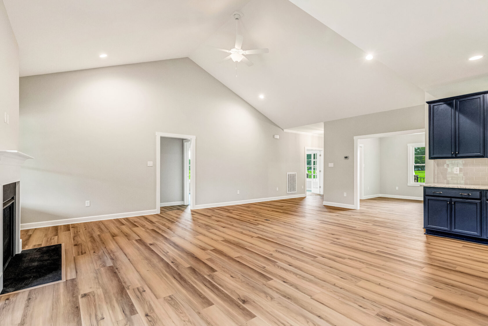Spacious, empty living room with wood floors, vaulted ceiling, fireplace, and view of kitchen and doorway.