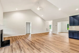 Spacious, empty living room with wood floors, vaulted ceiling, fireplace, and view of kitchen and doorway.