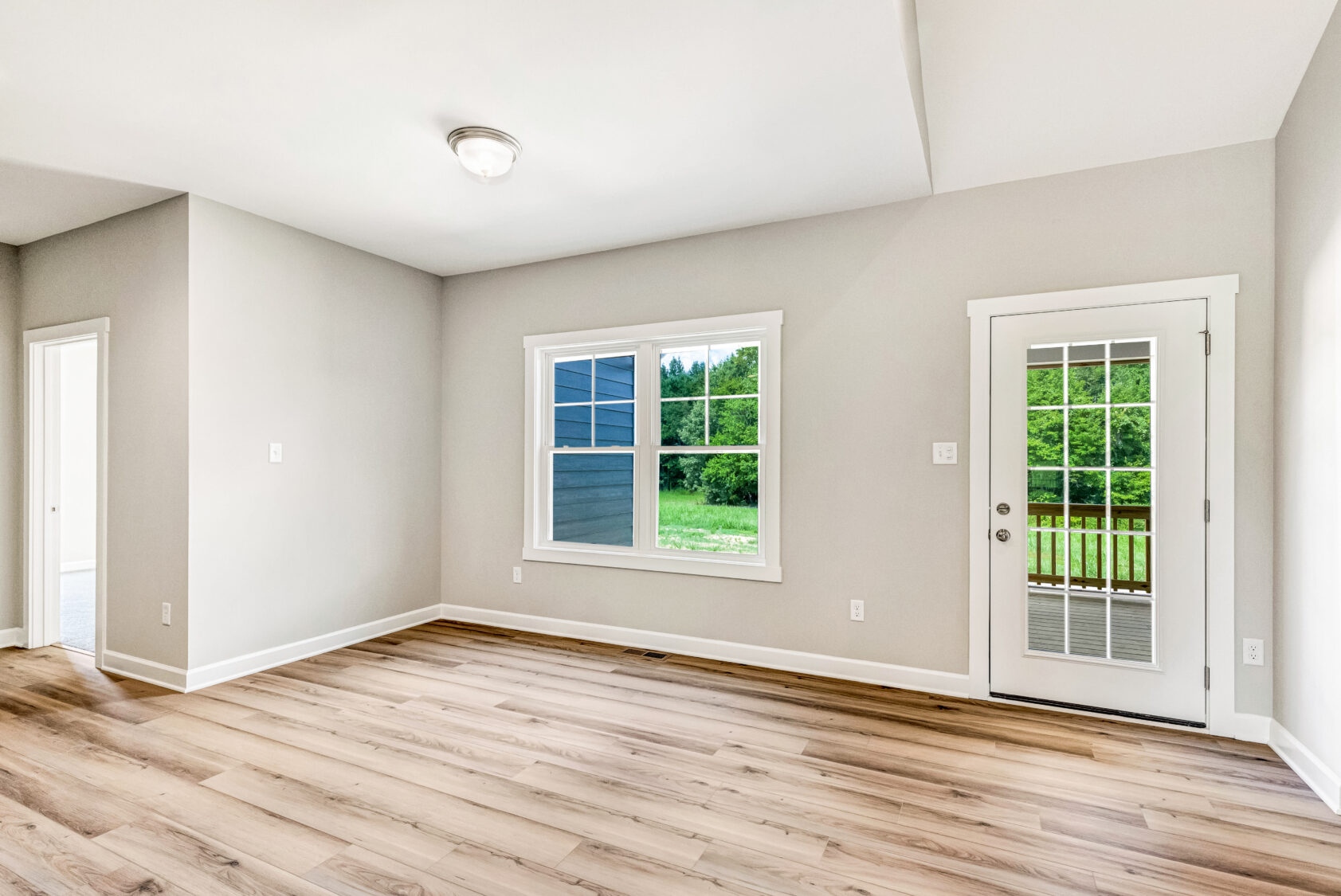 Bright empty room with light wood floors, beige walls, a large window, and a glass door to a green backyard.