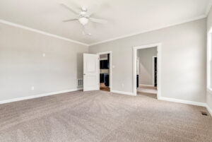 Empty carpeted room with light gray walls, ceiling fan, two open doors, and large window with natural light.