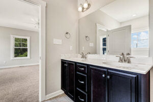 Modern bathroom with double sinks, dark cabinets, and a mirror, adjacent to a carpeted bedroom.