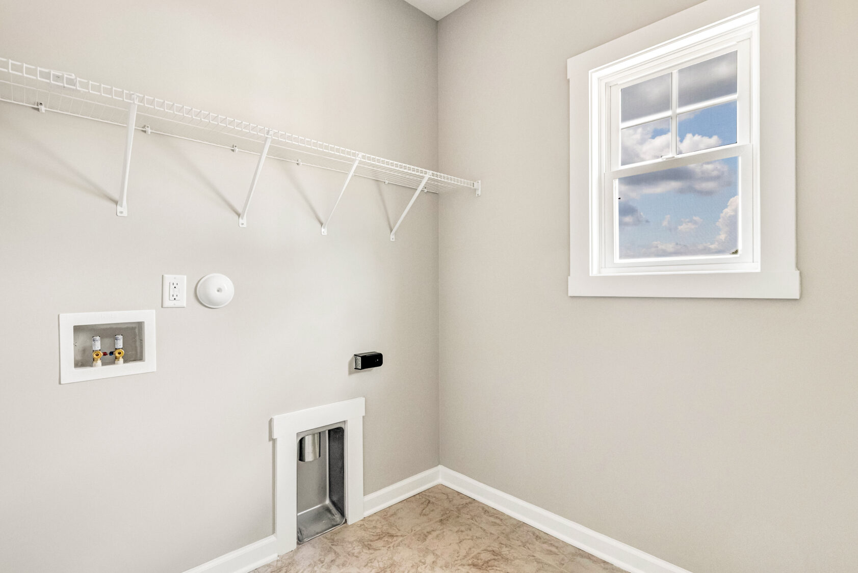 Laundry room with beige walls, empty wire shelf, washer hookups, and a window showing blue sky with clouds.